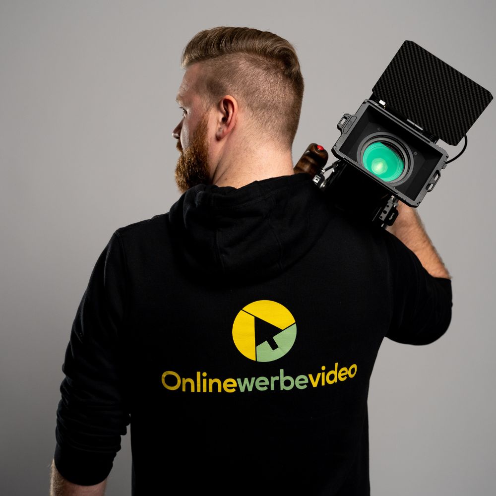 Videoproduktion Dresden Onlinewerbevideo zeigt einen professionellen Videomitarbeiter Philipp Züchner einer Videoproduktionsfirma, der mit dem Rücken zur Kamera steht und eine große professionelle Kamera hält, auf seinem Pullover steht Online Werbevideo Drehszenen, authentische Aufnahmesituation im Kundenunternehmen