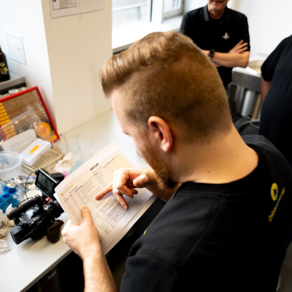 Imagefilm Drehvorbereitung mit Filmproduktionsmitarbeiter, der von hinten gefilmt Produktionspläne prüft, während eine Kamera im Vordergrund auf den Kundendreh ausgerichtet ist